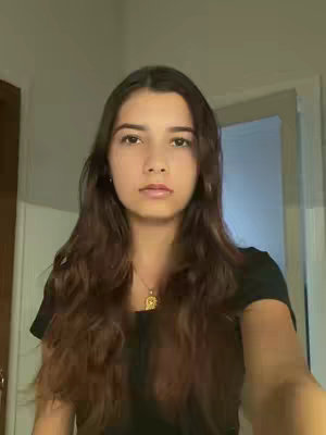 A young woman with long, wavy brown hair is centered in a well-lit room. She's wearing a black top and a necklace with a gold pendant. The background includes neutral-colored walls and slightly out-of-focus doorways or frames. Her expression is neutral, and her gaze is direct towards the camera.