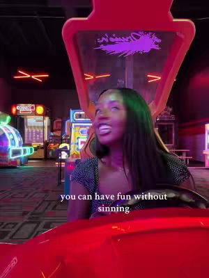 A woman is shown in an arcade setting, seated in a red race car game. She has long dark hair, and is looking upward with an excited expression. The arcade in the background is lit with colorful neon signs.