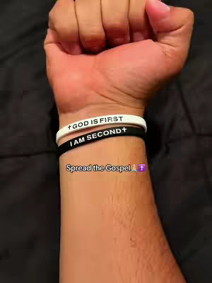 The image shows a close-up of a person's wrist and forearm, focusing on two stacked bracelets. The top bracelet is white with the text 'GOD IS FIRST' in black, and a small cross symbol at the beginning of the text. The bottom bracelet is black with the text 'I AM SECOND' in white, and a small cross symbol at the end of the text. Below the bracelets are the words 'Spread the Gospel' and a purple cross symbol.