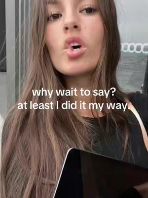 A young woman is shown from the chest up, looking directly at the camera with her mouth slightly open. Overlayed text in a white sans-serif font reads: 'why wait to say? at least I did it my way.' She is holding a black laptop or tablet in the foreground, and her brown hair is long and wavy, framing her face.