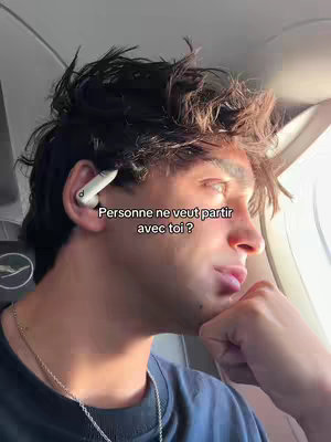 A young man looks out of an airplane window, resting his chin on his hand. Text overlay is placed in the top right corner of the frame. He has dark, tousled hair, and is wearing an ear bud with a dangling silver necklace with a black shirt. The background is partly obscured by the plane's wing or engine.