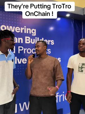 Three men are gathered in front of a blue backdrop with white text. The central figure is holding a microphone, while the other two stand on either side, listening. The image is brightly lit, and the overall impression suggests a presentation or a promotional event.