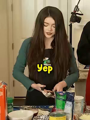 A young woman with long dark hair is shown in a kitchen setting, wearing a green shirt and a black apron with a small cartoon design. She is holding something between her hands near her waist, and a speech bubble with the word 'Yep' in yellow is overlaid near her midsection. Various kitchen items like bowls, pantry items, and a bag are visible in the background. She is looking down at what she is holding.