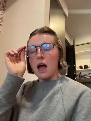 A woman with short hair and glasses looks at the camera with her mouth open and one hand touching her glasses. Her grey sweatshirt is visible.