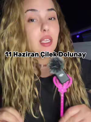 A woman with curly blonde hair is centered in the frame. She appears to be speaking, with her lips parted, and a microphone is held in front of her. The background is dark, with the focus on her face and upper body. The text overlay at the bottom reads in Turkish characters.