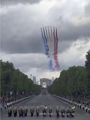 An exterior shot of a wide, tree lined avenue with the Arc de Triomphe in the background. Above the avenue there are colored smoke trails with the colors of the french flag. In the foreground, a marching band is on the lower center, followed by people walking in military uniforms. The sky is a cloudy grey.