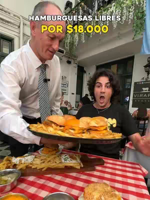 The cover image shows a restaurant scene with two men. The man on the left, dressed in formal attire, is holding a large platter of food. The man on the right is reacting to the food with an open-mouthed expression, wearing a black t-shirt. The scene is brightly lit, with a red and white checkered tablecloth in the foreground and a blurred restaurant setting in the background, the text overlay at the top announces "HAMBURGUESAS LIBRES POR $18.000".