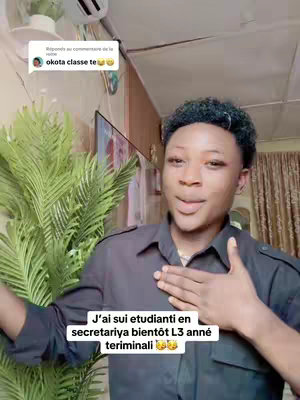 A young man with dark hair and makeup is shown, wearing a dark gray button-up shirt. He has a hand over his chest and is looking towards the camera. The background includes interior elements such as indoor plants in pots. At the top-left, a comment from a social media post is visible.