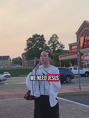 A woman with dark hair, wearing a white top, and a black skirt, stands on a brick surface in front of a street with cars and buildings. She is holding a microphone and speaking. The text "WE NEED JESUS" is overlaid on the image within a red rectangle.