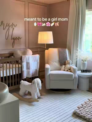 A well-lit, serene nursery in soft neutral tones. The room is decorated with a crib, a comfy armchair, and a rocking animal. The walls feature a decorative carved name, and the overall atmosphere suggests a baby's room.