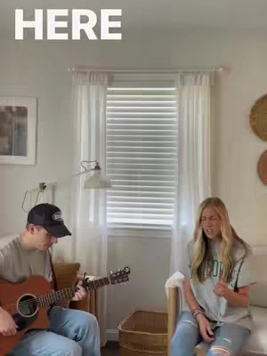 Two people, a man and a woman, are inside a room. The man on the left is sitting on a chair with a guitar, and is wearing a black hat. The woman is on the right, singing. The environment is a bright, well-lit room with a window.