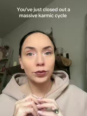 A woman with fair skin is centered in a selfie-style shot. Her dark hair is pulled back, and she wears a light-colored hoodie. The background includes a shelf and a glimpse of a mirror. Text at the top reads, 'You've just closed out a massive karmic cycle'.