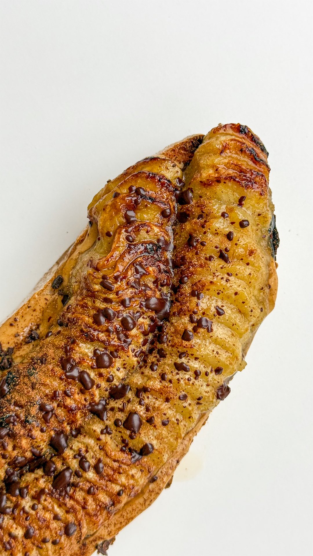 A close-up shot of a piece of food angled slightly to the upper right. The food has a golden-brown exterior, and a darker, possibly charred area near the top. Small, dark brown chocolate chips have been scattered across the surface. The background is plain white.
