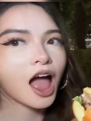 A close-up of a young woman's face with wide eyes and an open mouth, as if in surprise or anticipation. She is holding a burger, partially visible at the bottom right. The background is blurred, suggesting an outdoor setting at dusk.