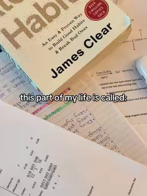 The image shows a desk covered in open books and papers, with a copy of "Atomic Habits" by James Clear prominently displayed. The book is beige with large text.  In the foreground study notes with highlighted texts and handwritten annotations with various diagrams and data is visible. The overall impression is one of focused study.