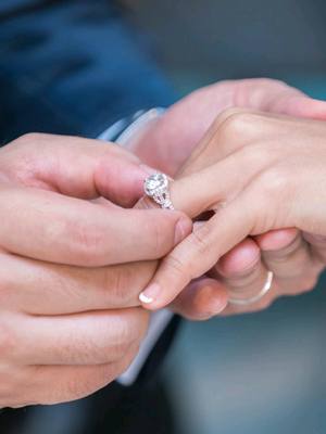 Close-up of a person placing a diamond engagement ring on another person's finger. The focus is on the hands and the ring. The background is blurred and out of focus.