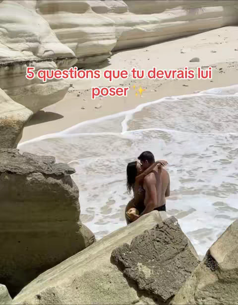 A couple kissing on a beach, embraced in front of rough white rock formations and ocean waves.