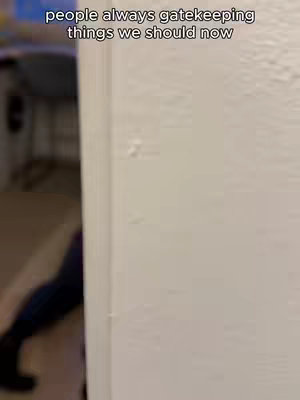 The image features a close-up of a white wall with a slightly open doorway on the left. The wall is textured, and there are a few minor imperfections visible. Through the doorway, a blurred figure is visible, suggesting movement or a person partially in view.