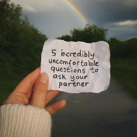 A hand holding a crumpled piece of paper with handwritten text against a backdrop of a road, trees, and a rainbow. The paper is centered and the fingers are holding it up, with the background slightly blurred.