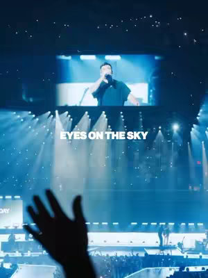 The image shows a live concert setting with a singer on a lit-up stage, a raised hand in silhouette, and the words "EYES ON THE SKY" superimposed. The dominant colors are shades of blue and white, creating a dynamic and spiritual atmosphere.