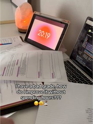 The image displays a study setup on a wooden desk. A laptop is open on the right side, showing a screen with elements of a periodic table and a bar. In the center, an open tablet displaying a timer that reads "20:19" is the primary focus. The background includes a glowing pink salt lamp. Scattered printed notes and pages cover the desktop, with text that suggests study notes.