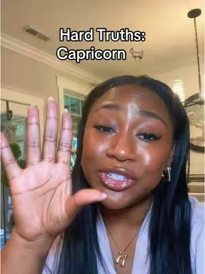 The image shows a close-up of a Black woman. The woman has dark hair and is wearing a light-colored top with a gold necklace. She is holding up her right hand with her fingers spread. Above her head, the text "Hard Truths: Capricorn" is displayed in a white font. The background appears to be a home interior, with a light fixture and portions of a window visible. Her facial expression appears to be of surprise or storytelling, with her lips slightly parted and looking towards the camera.