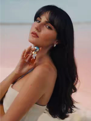 A woman with long dark hair and bangs is the central figure in a photograph. She is wearing a white strapless top and elegant earrings. Her gaze is directed slightly upwards, and she gently touches her neck with one hand. The background is a soft blend of pink and blue.
