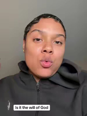 A close-up shot of a woman with short, styled hair wearing a black zip-up hoodie. She is looking directly at the camera with a neutral expression. The background is a plain, light gray wall. The composition is simple, focusing on the woman's face and upper body.
