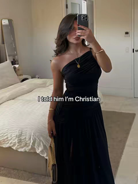A woman in a black one-shoulder dress is taking a selfie in a bedroom. She is holding a phone to take the selfie, her face is partially obscured. She has long, dark hair styled with soft layers. She is wearing jewelry and has a small gold purse. In the background is a made bed, mirror and door.