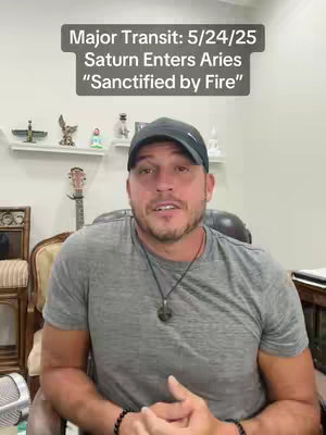 The cover image features a man in a casual setting, presumably indoors. He is wearing a dark baseball cap and a gray t-shirt. The background includes a variety of decorative items on a shelf, a guitar leaned against the wall, and furniture. A text overlay at the top indicates the video content relates to a major transit, Saturn entering Aries, and the phrase "Sanctified by Fire". The man is looking directly at the camera with a neutral expression.