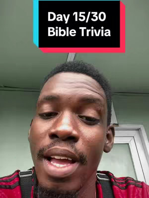 A selfie-style video cover image shows a Black man from the chest up, set against a light green wall. He gazes at the camera with a slightly open mouth, possibly speaking. Above his head, a rectangular text overlay in white font on a background, divided into teal and black. The text says "Day 15/30 Bible Trivia". He wears a red shirt with black arm stripes and a black shoulder strap.