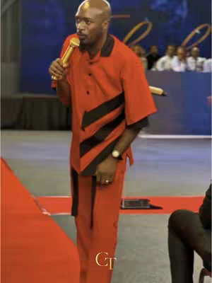 A man in a red and black suit stands on a stage with a gold microphone. He has a bald head and is speaking. Behind him, there is a dark blue backdrop with golden swirls and text that cannot be seen. In the bottom right corner, there is a 'GT' logo.