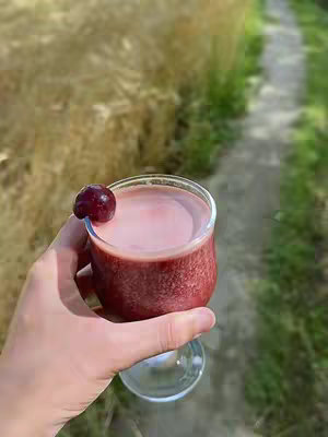 A hand holding a clear glass of a reddish-pink drink, garnished with a dark cherry. The background is blurred, showing a pathway and grassy areas.