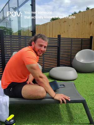 A man sits on a lounge chair outdoors, smiling at the camera. He wears an orange t-shirt and black shorts. The background includes a fence, a patio, and a small grassy area.