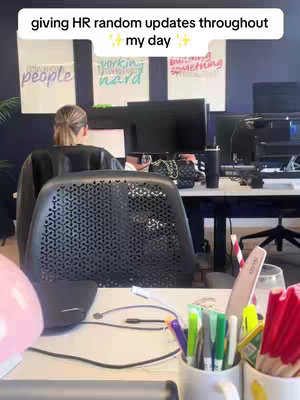 The image shows a woman from the back, working in what appears to be an office setting. The layout includes a desk with office supplies, computer monitors, and inspiring posters.