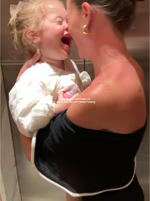 A woman holds a small child close, both laughing. The woman has her back to the camera, dressed in a black off-the-shoulder top. The child wears a white outfit with a bow in their hair.