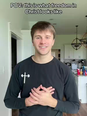 A man with short, brown hair is centered in the frame, wearing a black long-sleeved shirt with a white cross design. He smiles warmly with his hands clasped over his heart. The background shows a light-colored interior, with a door and kitchen visible, lit by natural light.