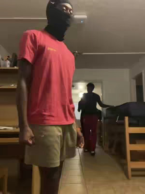 A person wearing a black balaclava and a red t-shirt stands in a room with two other people in the background. The person in the foreground is positioned left of the center. A shelf is visible on the left side. A door with light illuminating the hallway is visible on the right. Light from the ceiling is reflected on the ceiling.