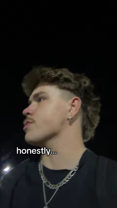 A young man with a mullet hairstyle and a silver chain necklace is shown in a close-up shot. He's looking up and to the left, with a slightly shadowed face and a dark background.