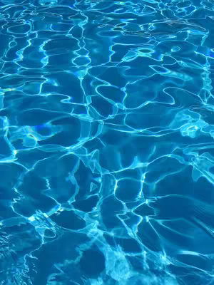 An abstract image of water, likely a pool or the sea. The water is a vibrant shade of blue, with sunlight reflecting off the surface creating bright, wavy patterns and highlights. The overall look is fluid and dynamic, with light and shadow shifting across the surface.