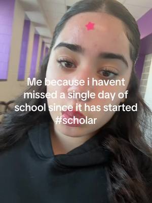 A selfie-style image with a young woman in the foreground. She is looking slightly down at the camera with a neutral expression. She has dark hair, eyebrows, and lashes. A small pink star sticker is placed on her forehead. The background shows a corridor with white and purple walls. Text is overlaid on the image.