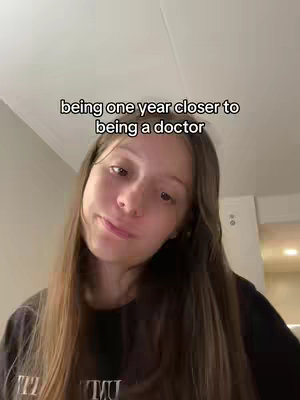 A young woman with long brown hair is in a selfie shot. The text overlay reads, 'being one year closer to being a doctor.' The background is a light-colored wall and some of the room is visible. The woman is looking at the camera, with a slightly upward head tilt, a soft expression, and looking towards the camera. She is wearing a dark shirt.