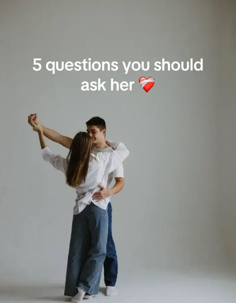 A couple are dancing in front of a plain gray background. The man is behind the woman with his arms wrapped around her waist. The woman's arms are raised and her head is turned towards the man. Text overlay at the top says, '5 questions you should ask her' followed by a heart icon.