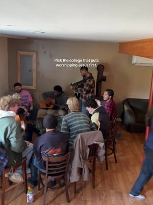 A group of people are gathered in a room with wood flooring, playing music and singing. The lighting is warm. There is a wooden cross.