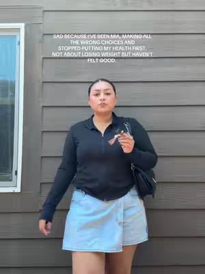 A woman stands in front of a gray wooden wall, wearing a black zip-up top and a light blue skirt. Text overlay in white font.