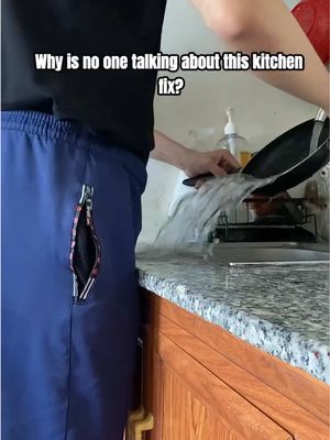 An individual pouring water from a pan into a sink. The person is wearing dark shorts with a zipped pocket. The countertop is made of marble. Above, a white text overlay asks, "Why is no one talking about this kitchen fix?"