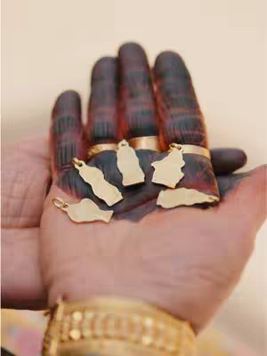 A close-up shot of a person's hand, heavily adorned with henna or a similar dark dye. The hand is open, and several gold charms, each shaped like a different Middle Eastern country, rest in the palm and attached to rings on the fingers. A gold bracelet is visible around the wrist, and the backdrop is a soft, neutral tone, likely a light beige or tan.
