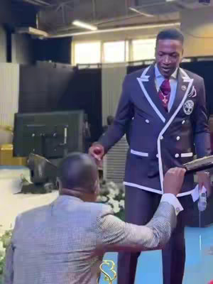 The image shows a well-lit indoor scene with two men, one in a light-colored suit kneeling, and the other in a dark suit standing above, holding his hand out. The composition emphasizes a moment of interaction between them, possibly a moment of receiving guidance.