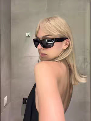 A woman with blonde hair wearing sunglasses and a black top stands in front of a light gray tiled wall. The image is taken from a slightly low angle, with the wall tiles and the woman's silhouette occupying most of the frame. The woman has blonde hair, cut with a long fringe and is wearing black sunglasses. She is looking over her shoulder, and is only partially visible, revealing her bare back. The lighting seems to be natural with a soft reflection.