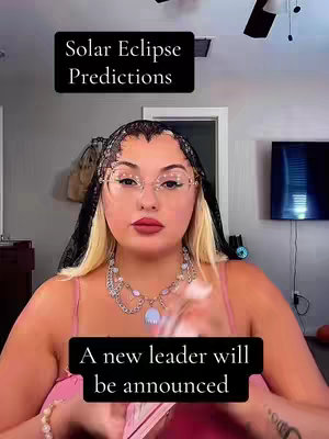 A woman is centered in the frame. She has blonde and black hair, is wearing glasses and a necklace with a blue center piece. She is wearing a pink top. The background has a fan, a dark wall and a door. There are two black rectangles with the text 'Solar Eclipse Predictions' and 'A new leader will be announced'.